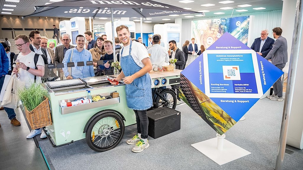 Ein Eismann mit blauer Schürze lächelt und verteilt Hörnchen mit Eis an die Messebesucher, daneben ein Infowürfel. Im Hintergrund stehen Personen um das Eisfahrrad mit Anhänger an einem Stand auf der polisMOBILITY in Köln. 