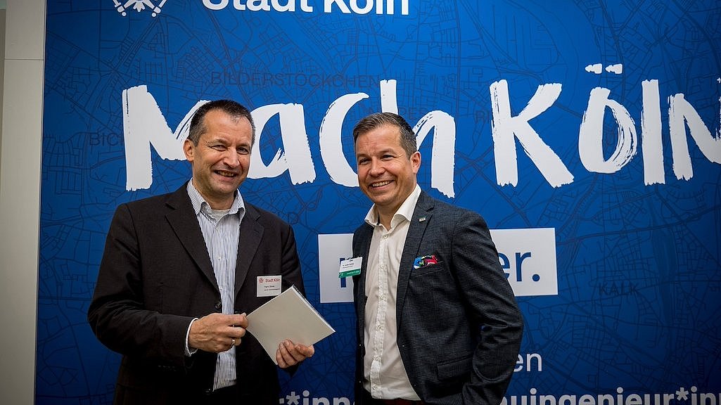 Zwei Herren (Patric Stieler und Dr. Johannes Harding) vor einem blauen Plakat mit der Aufschrift Stadt Köln - Mach Köln auf der Kongressmesse polisMOBILITY. 