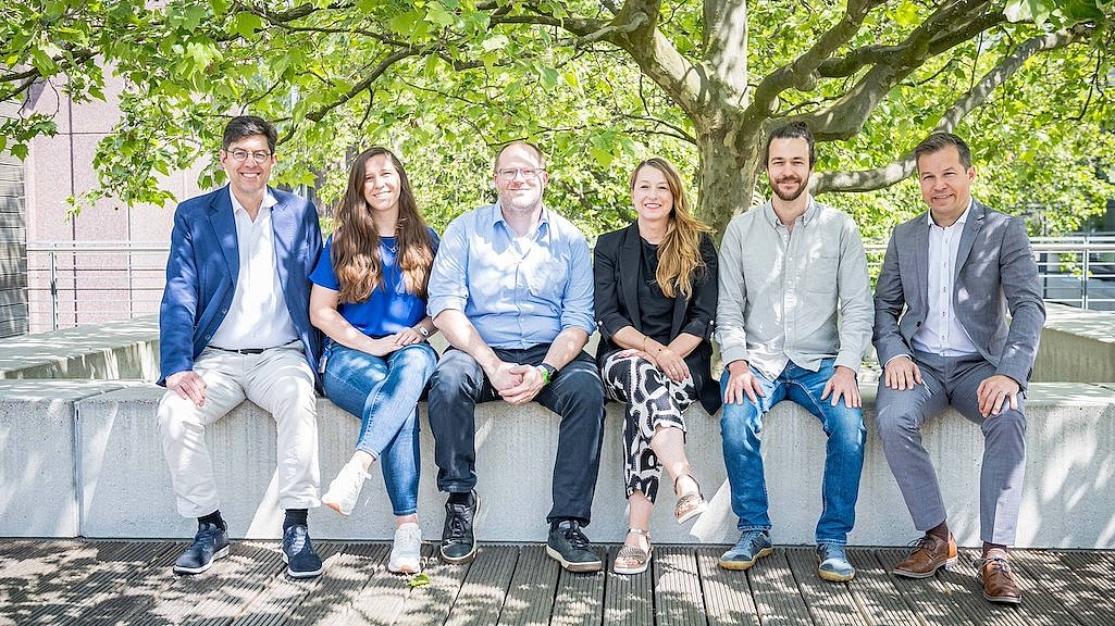 Gruppe von sechs Menschen draußen im Halbschatten auf einer hohen Umrandung in Reihe vor einem Baum sitzend (Harald Fletcher, Lars Röglin, Margot Dittmann und Dr. Jochen Harding vom Mobidrom und Anna Beer sowie Bennedikt Spitzley vom Team SEVAS).