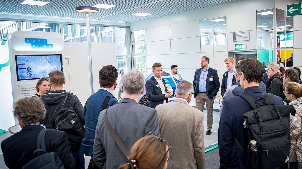 Eine Gruppe von Menschen steht an einem Messestand des Mobidroms auf der polisMOBILITY und horcht einem Vortrag. Im Hintergrund ein großer Bildschirm.