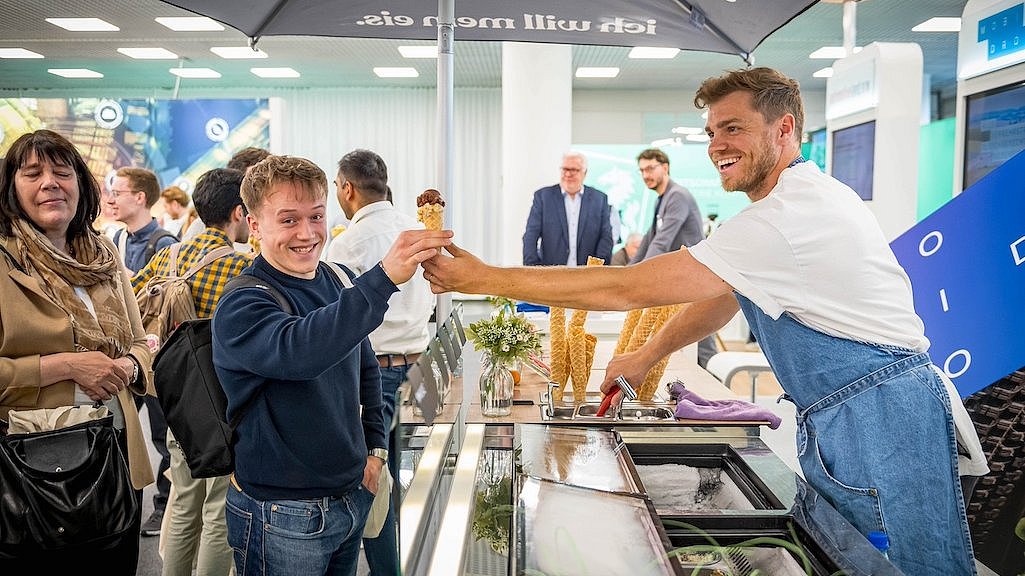 Menschen stehen um ein Eisfahrrad mit Anhänger an einem Stand auf der polisMOBILITY in Köln. Der Eismann gibt ein Hörnchen mit Eis an einen lächelnden Messebesucher aus.