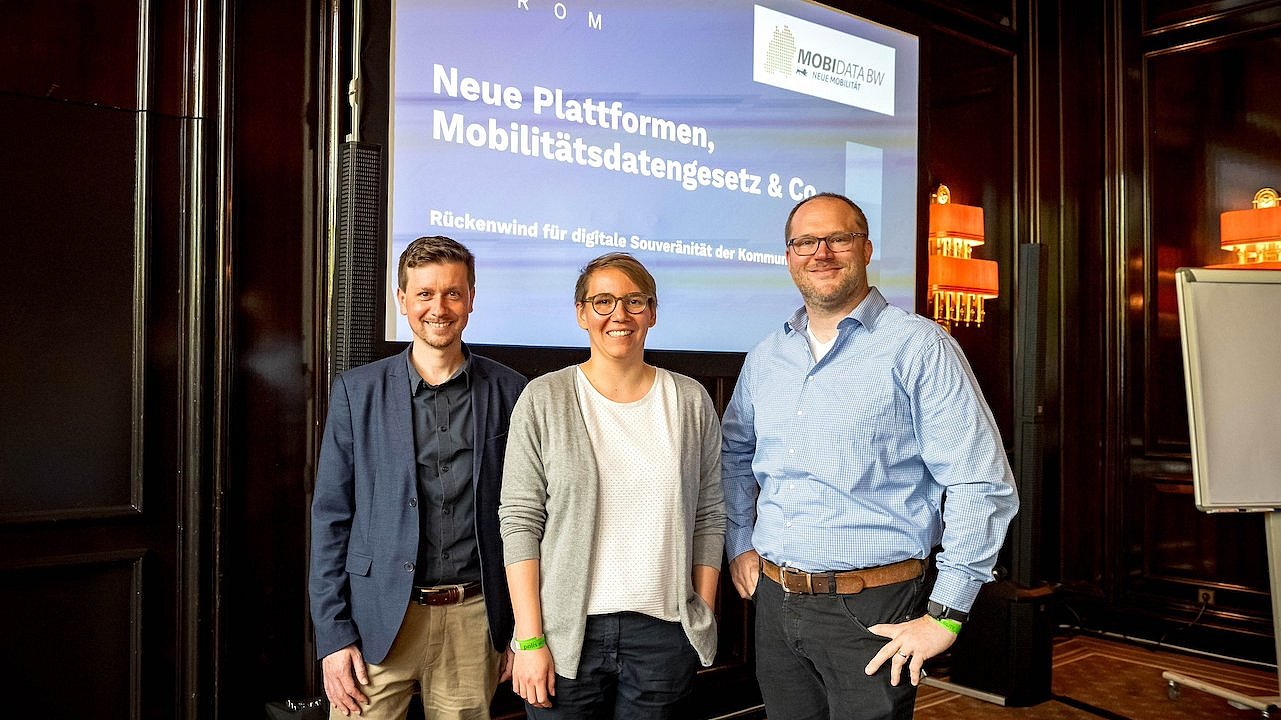 Drei Redner vor einem großen Bildschirm auf einem Workshop auf der polisMOBILITY in Köln (Marlene Picha, Innovationsmanagerin von MobiData BW, Dr. Roland Goetzke, Bundesministerium für Digitales und Verkehr und Lars Röglin, Mobidrom).