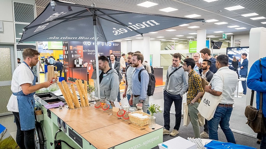 Menschen stehen um ein Eisfahrrad mit Anhänger an einem Stand auf der polisMOBILITY in Köln. Es werden Hörnchen mit Eis an die Messebesucher verteilt. 